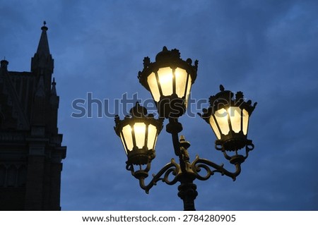 Gothic lamppost with lit lanterns illuminates a historic urban street at dusk with soft warm glow and quiet night atmosphere