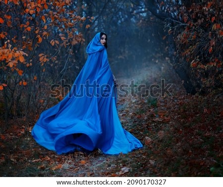 Gothic mysterious woman walks in fantasy autumn forest. long silk blue cloak flutters, waving fly in wind, fabric in motion. Head hood. Girl princess looks back. Fallen orange leaves dark trees, fog
