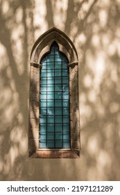 A Gothic Style Church Window With Iron Bars With A Green Curtain And A Tree’s Shadow.