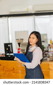 Gotpam Asian Female Barista In Front Of The Shop. A Successful Small Business Owner Smiles And Stands In Front Of The Counter Wearing An Apron Holding A Tablet In Front Of A Coffee Shop Counter.