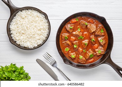Goulash traditionellen ungarischen Rindfleisch Eintopf Suppe Essen Abendessen gekochtes Rezept mit würziger Gravy-Sauce in Gusseisen Pfanne serviert mit Reis und zerkleinerte Petersilie auf weißem Holztisch-Hintergrund.