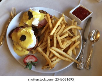 A gourmet breakfast plate featuring eggs benedict with truffle garnish, seasoned fries, and a side of ketchup, elegantly presented with silverware on a white tablecloth - Powered by Shutterstock