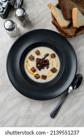 Gourmet Cream Soup In A Matt Black Bowl, Above View, On White Marble Table With Bead Basket Aside