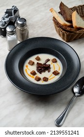 Gourmet Cream Soup In A Matt Black Bowl, Above View, On White Marble Table With Bead Basket Aside