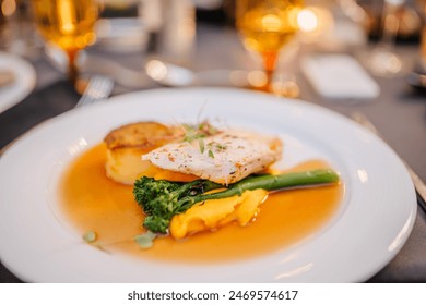 Gourmet dish with fish fillet, roasted potatoes, broccolini, and sauce, elegantly plated on a white dish with a blurred background of dining setting. - Powered by Shutterstock