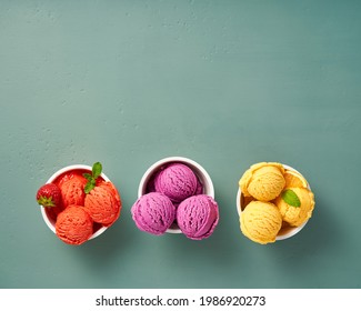 Gourmet Flavours Of Italian Ice Cream In Vibrant Colors Served In Individual Paper Cups On Blue Background. Strawberry Sorbet, Blueberry Or Lavender Ice Cream With Orange Sorbet Decoration With Mint