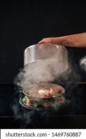 Gourmet Food. Hand Holding Cloche Over Burnt Caprese Salad On A Dark Background.