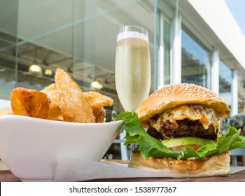 Gourmet Hamburger With Melted Cheese, Avocado, Bacon, Lettuce, Tomato And Salad Sauce On A Sesame Seed Bun Plus Side Serving Of Crisp Fried Potato Wedges And A Glass Of Sparkling Wine Or Champagne 