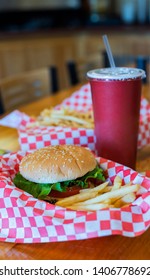 Gourmet Hamburger On Red Checkered Paper At Restaurant On Table With Fries, Dipping Sauce, And A To Go Soda Cup