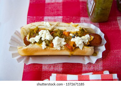 Gourmet Hotdog With Feta Cheese, Roasted Peppers And Pickle Relish Sitting In Paper Holder On Red Checkerboard Tablecloth With Relish Bottle - 4th Of July BBQ Food