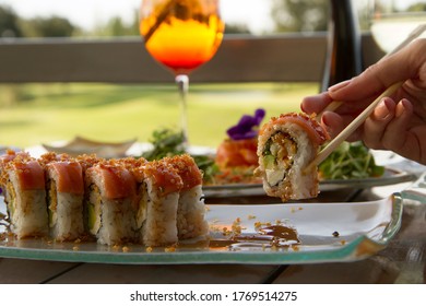 Gourmet Seafood. Elegant Sushi Restaurant. Dish Presentation. Young Woman Dining Outside With A Beautiful Natural Background. Close Up Of A Woman Hand Holding Chopsticks With A Raw Salmon Sushi Roll. 