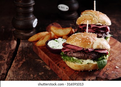 Gourmet Tasty Steak Burgers with Ham Slices on a Wooden Tray with Potato Wedges and Dipping Sauce.