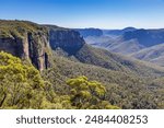 Govetts Leap Lookout, a panoramic view of the Blue Mountains National Park in New South Wales, Australia, featuring a lush valley, rugged cliffs, and a clear blue sky.