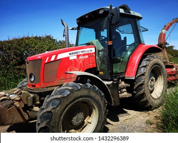 Gower, UK: June 23, 2019: A Farm Worker Is Driving A Tractor In A Lane With High Hedges Around The Edge Of A Field. Agricultural Machinery At Work. 