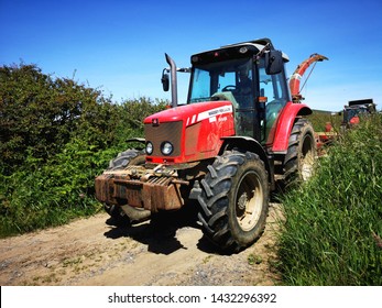 Gower, UK: June 23, 2019: A Farm Worker Is Driving A Tractor In A Lane With High Hedges Around The Edge Of A Field. Agricultural Machinery At Work. 