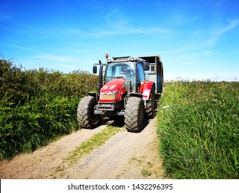 Gower, UK: June 23, 2019: A Farm Worker Is Driving A Tractor In A Lane With High Hedges Around The Edge Of A Field. Agricultural Machinery At Work. 