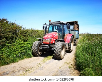 Gower, UK: June 23, 2019: A Farm Worker Is Driving A Tractor In A Lane With High Hedges Around The Edge Of A Field. Agricultural Machinery At Work. 