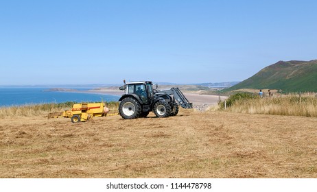 Gower, Wales, UK: July 26, 2018: A Tractor Pulling Agricultural Machinery Bales Straw While Farm Workers Clear The Bales At Rhossili On The Gower Peninsular In A Panoramic Format.