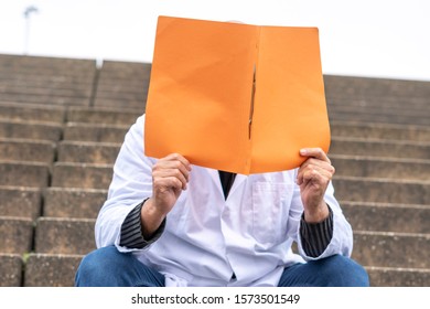 GP Medical Doctor Or Dentist Sitting On Outdoor Stairs And Hiding His Face With A Clinical Study Report