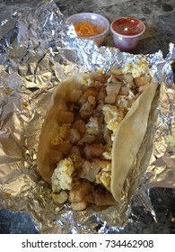 Grab A Austin , Texas Tex-mex Flour Tortilla BreakFast Taco With Scrambled Eggs And Potatoes  On Foil With Cheese And Salsa In The Background 