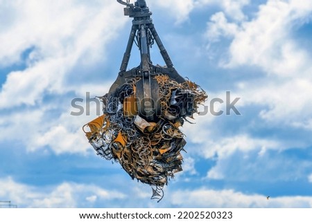 Grab crane loading iron and steel waste, scrap metal for recycling at a landfill in an industrial waste collection center with small falling particles of metal chips and dust.