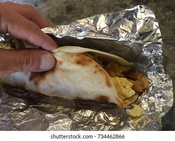 Grabbing A Austin , Texas Tex-mex Flour Tortilla BreakFast Taco With Scrambled Eggs And Bacon And Potatoes  On Foil 