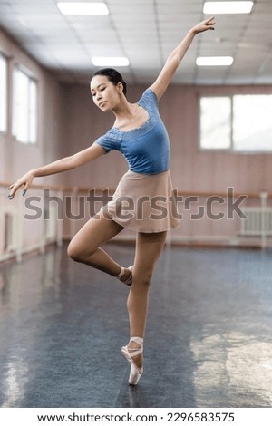 Graceful Asian ballerina in a blue bodysuit and a beige skirt is rehearsing in a dance class. 