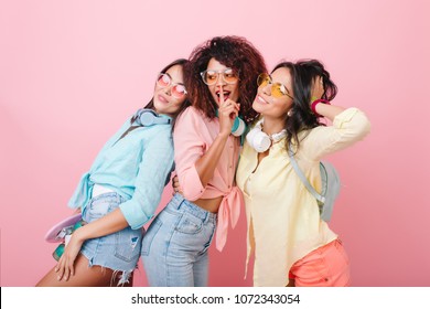 Graceful Asian Girl With Backpack Posing Beside Friends During Joint Photoshoot In Pink Studio. Indoor Photo Of African Glamorous Lady Telling Secret To Brunette Female Models.