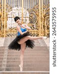 Graceful ballerina in black tutu and blue leotard posing en pointe on stone steps in front of ornate golden gate, performing an elegant dance move outdoors in Paris.