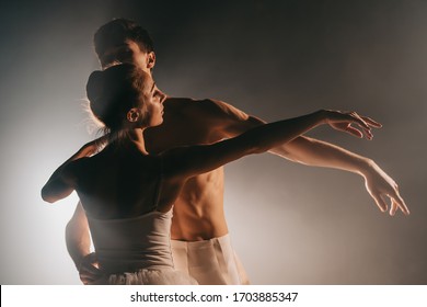 Graceful Ballerina And Her Male Partner Dancing Elements Of Classical Or Modern Ballet In Dark With Floodlight Backlight. Couple In Smoke On Black Background. Art Concept.