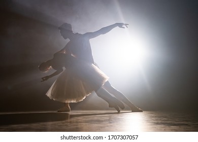 Graceful Ballerina And Her Male Partner Dancing Elements Of Classical Or Modern Ballet In Dark With Floodlight Backlight. Couple In Smoke On Black Background. Art Concept.