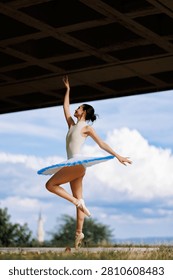 Graceful ballerina performs an elevated pose on pointe outdoors beneath an overpass, framed by blue sky and clouds.