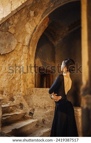 Graceful ballerina is standing on old rustic staircase of a damaged building wearing elegant black dress and leotard with arms around her waist. Chic ballet diva wearing dress poses in ruined building