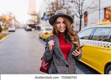 Graceful brunette woman in vintage coat walking down the street with phone in hand. Outdoor portrait of smiling refined girl in hat an red sweater standing next to yellow taxi. - Powered by Shutterstock