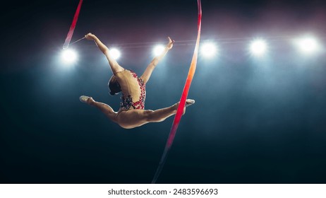 Graceful Female Gymnast in Mid-air, Performing Split Leap With Ribbon Under Bright Stadium Lights. Flexibility, Elegance, and Precision in Captivating Rhythmic Gymnastics Routine. - Powered by Shutterstock