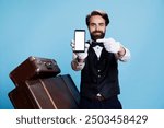 Graceful hotel porter posing next to stack of suitcases while using device with white screen. Presenting blank smartphone display, bellhop illustrates dedication of his line of work.