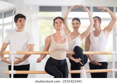 Graceful positive woman, instructor of ballet class for adults, posing with students at barre during rehearsal break in light-filled choreography studio - Powered by Shutterstock