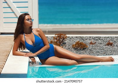 Graceful tanned woman with long legs in sunglasses and blue one-piece swimsuit lies taking sunbathe near water pool at tropical resort hotel