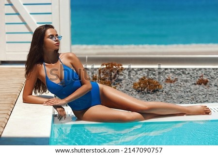 Graceful tanned woman with long legs in sunglasses and blue one-piece swimsuit lies taking sunbathe near water pool at tropical resort hotel