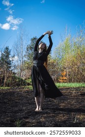 Graceful witch woman in a black dress dancing on burnt grass outdoors in a forest with sunlight and trees. Concept of nature recovery.