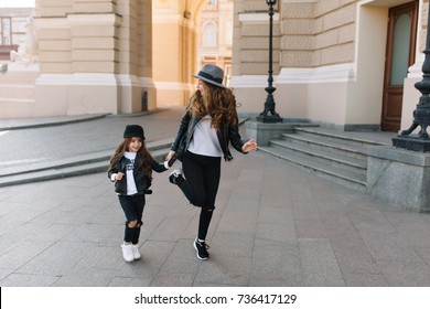 Graceful Young Curly Woman In Black Skinny Jeans Funny Dancing On The Street Near The Little Joyful Girl. Portrait Of Stylish  Mom In Leather Jacket And Charming Daughter Having Fun Outside