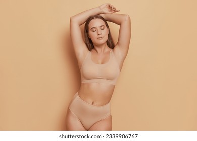 Graceful young woman wearing beige bra and panties stands at light brown background, hands over her head, natural beauty concept, copy space