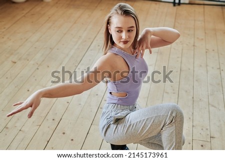 Gracious teenage girl in activewear outstretching arm while doing exercise during vogue dance training on wooden floor of modern studio