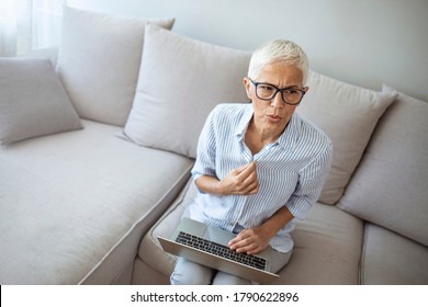 Gradual Process.Beautiful Mature Woman Touching Shirt And Having Hot Flash. Exhausted Mature Woman Entering Menopause. Portrait Of An Attractive Senior Woman Sitting On A Sofa At Home With A Hot Flash