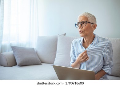 Gradual Process.Beautiful Mature Woman Touching Shirt And Having Hot Flash. Exhausted Mature Woman Entering Menopause. Portrait Of An Attractive Senior Woman Sitting On A Sofa At Home With A Hot Flash