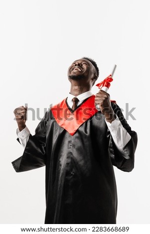 Graduate african man is graduating college and celebrating academic achievement. Happy african student in black graduation gown and cap raises masters degree diploma above head on white background