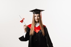 Graduate Girl With Master Degree Diploma In Black Graduation Gown And Cap On White Background. Happy Young Woman Successfully Graduated From The University With Honors