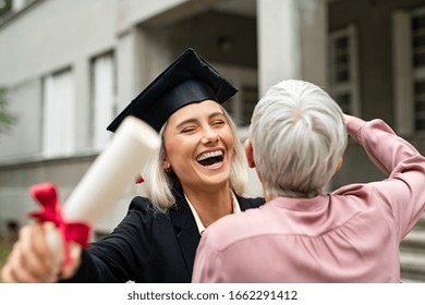Graduate student in black gown wearing mortar board and holding degree running to embrace mother in campus. Portrait of excited young woman hugging her mom at graduation ceremony in university.
