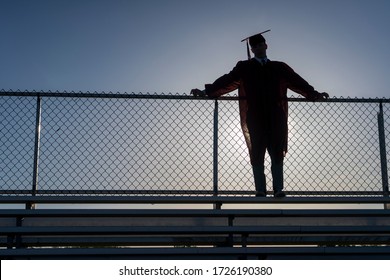 A graduating high school teenage boy wearing a cap and gown leans against a stadium fence and is silhouetted by the setting sun.  - Powered by Shutterstock