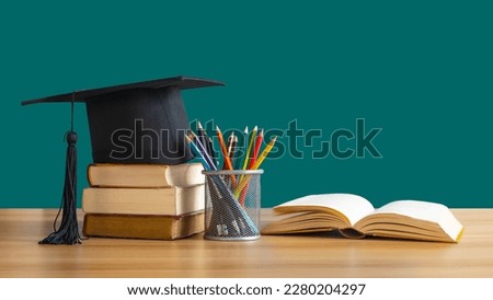 A Graduation cap placed on a stack of books with pencils next to it and an open book on the table. It is the concept of education.